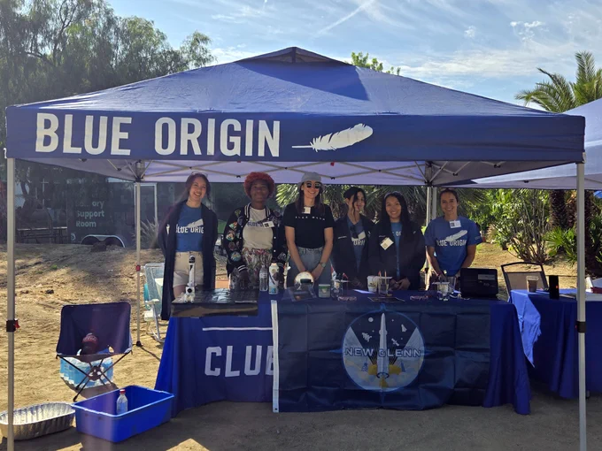 A group of people stand under a blue tent with the words "Blue Origin." The tent shades a table displaying various items and promotional materials. Several people are wearing matching shirts, and there is a banner on the table with the New Glenn logo.