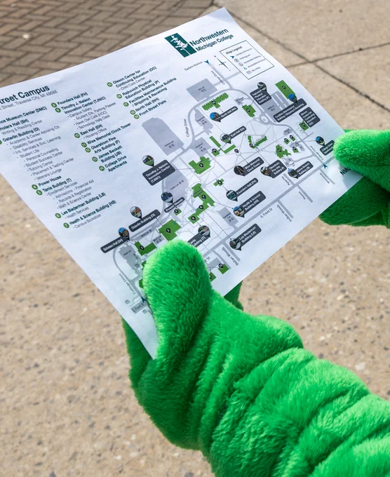 A person in a bright green, fuzzy costume is holding a detailed map of the Northwestern University campus. The map includes various buildings and pathways marked with symbols and text. The background shows a paved surface.