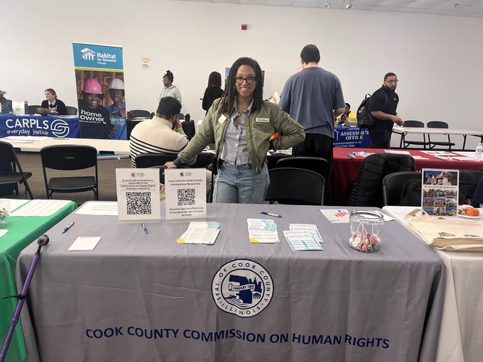 An individual wearing a green jacket, button up blouse, and glasses stands behind a table. On the table there is a branded Cook County Commission on Human Rights tablecloth. The table also includes paper handouts, pens, and candy.