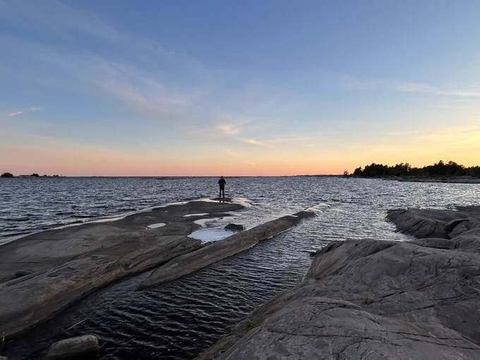 Person standing on a rocky shoreline with a sunset behind them.