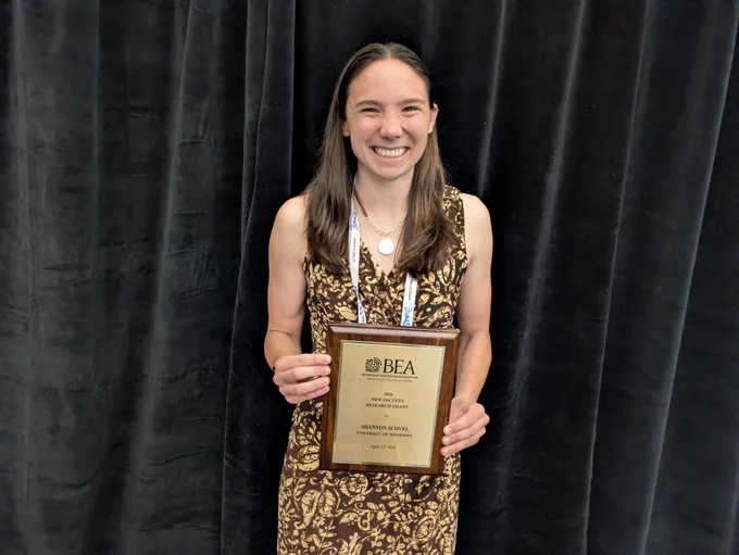 Photo of a woman holding a plaque in her hand. She is wearing a brown dress with a flower pattern on it and has brown hair. She has a gold necklace on and the plaque she is holding says B-E-A Broadcast Education Association 2026 new faculty research grant Shannon Scovel University of Tennessee April 17, 2026 on it.