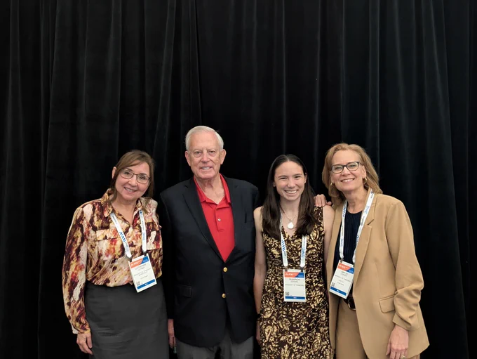Photo of four people standing posing for a photo in front of a black curtain. The woman on the far left has brown hair and is wearing a patterned shirt with black skirt with a lanyard on. The man next to her is wearing a black blazer with a red shirt and is smiling with gray hair. The woman next to him is wearing a brown dress with a flower pattern on it and has brown hair. She has a gold necklace on and is wearing a lanyard. The woman on the far right is wearing a brown suit with a lanyard on and brown hair and black framed glasses.