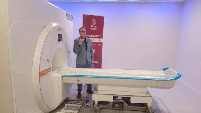 University of South Carolina Vice President for Research uses his hands to illustrate a point as he speaks with representatives of the media (just off camera) about USC's new 7-Tesla MRI scanner. He stands in front of a garnet "USC Brain Health" banner and just behind the beautiful new MRI scanner, which is white and still bears protective blue plastic used to protect the sensitive equipment during shipping and installation.