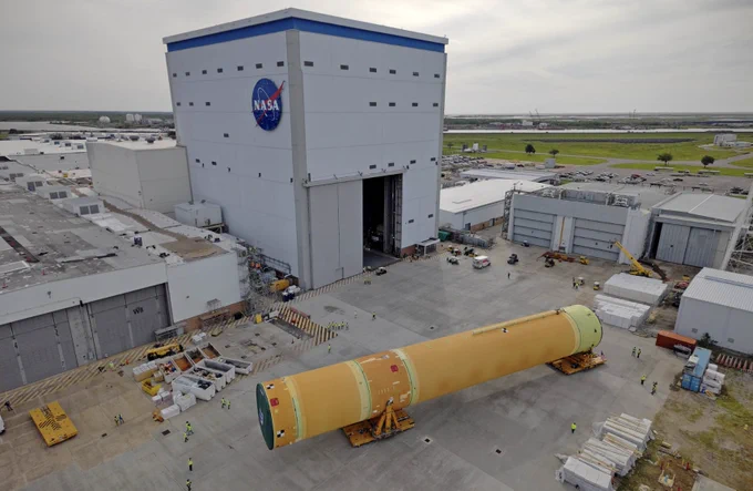NASA moved the core stage, or the largest section, of the SLS (Space Launch System) rocket that will launch the crewed Artemis III mission in 2027 from the agency's Michoud Assembly Facility to the agency\u2019s Pegasus barge in New Orleans on April 20, 2026. The core stage is a 212-feet long orange cylinder oriented on its side. The core stage is resting on stands in front of a large warehouse with a NASA logo on it. Credit: NASA/Michael DeMocker