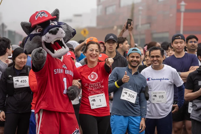 I am standing with Stony Brook University community members and Wolfie at the 5K run/walk outside of the Union Hall. an Some individuals have numbered race bibs, while others wear casual athletic attire. 
