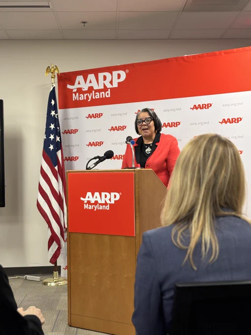 Here’s Acting Secretary Stacy Rodgers speaking at a podium during an AARP Maryland event. She is wearing a red blazer and standing in front of an AARP Maryland backdrop with an American flag to her left.