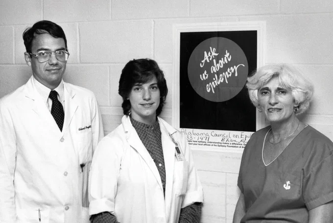 Black-and-white photograph of three medical staff members standing indoors against a cinderblock wall, two wearing white lab coats and one wearing medical scrubs. A poster on the wall reads, “Ask us about epilepsy,” with smaller text below referencing the Alabama Council on Epilepsy and a telephone number. The image appears to document an educational or awareness effort related to epilepsy.