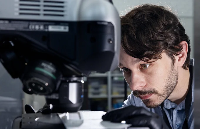 A person is intently examining a microscope in a laboratory setting. Shelves with various items are blurred in the background.