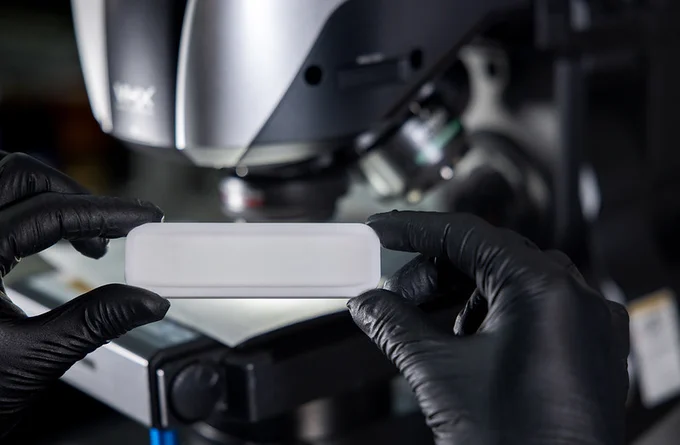 Two gloved hands holding a microscope slide in front of a microscope.