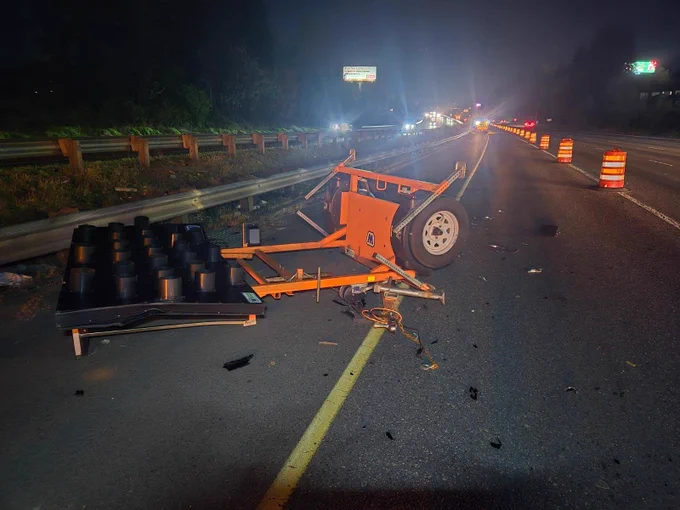 A portable electronic highway message board on wheels, crumpled and broken, laying face-down on the shoulder of a highway at night. Orange road barrels line the lane beside it.