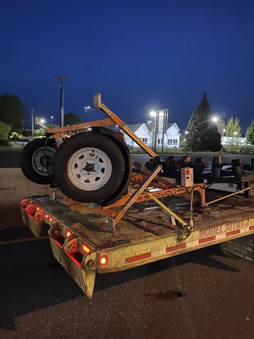 A portable electronic highway message board on wheels, crumpled and broken on the back of a flatbed truck, on the side of a highway at night.