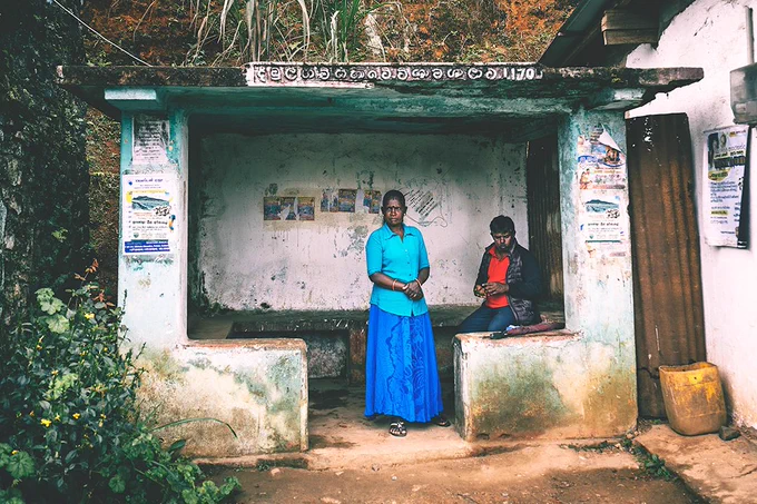 A Sri Lankan woman im a bright blue blouse and skirt stands inside a small bus shelter. A man in a bright red shirt seats next to her.