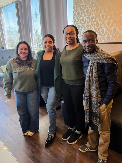 Four people standing indoors near large windows with natural light, dressed casually in sweaters and jackets, posing for a group photo.