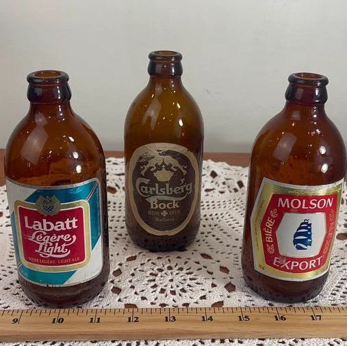 Three brown glass beer bottles placed on a lace doily atop a wooden surface. From left to right: a Labatt Lager Light bottle with a blue and red label, a Carlsberg Bock bottle with a dark label, and a Molson Export bottle with a red and gold label featuring a sailing ship. A ruler runs along the front edge of the table.
