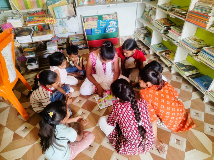 A group of young children sits in a circle on the floor of a colorful community library, gathered closely around a librarian who is reading a comic book aloud. Bookshelves filled with children’s books surround them, and the children lean in with focused, curious expressions as they follow the story together.