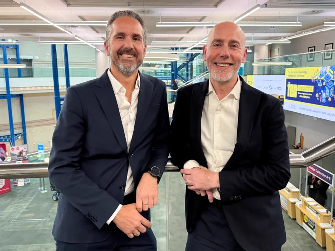 Two men in business suits standing inside a modern office building with glass railings and digital displays in the background. On the left is Geir Tore Jakobsen, the CEO of Hepro Group, and on the right is David Hammond, CEO of Chiptech UK and Europe.