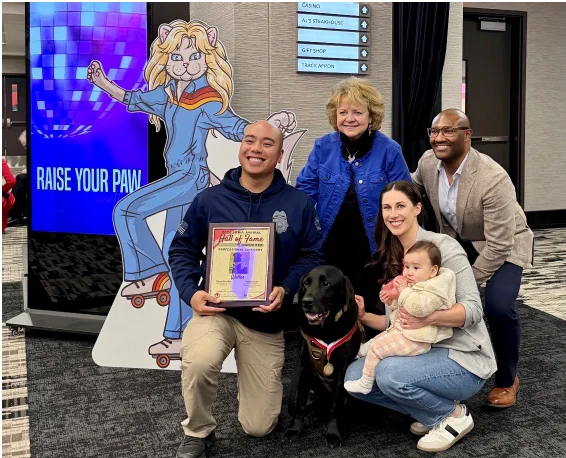Group photo with a man holding a framed award, a black dog wearing a medal, and family members posing indoors at a ceremony.