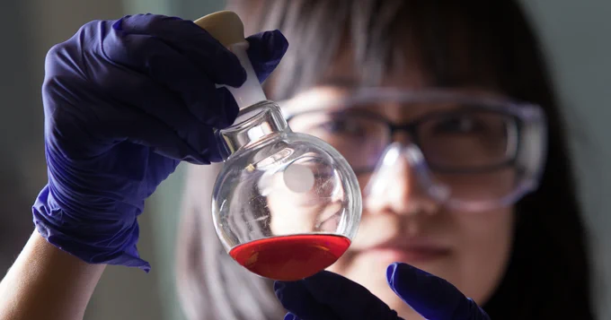 A person wearing safety goggles and blue gloves holds up a round-bottom flask containing a red liquid, inspecting it closely.