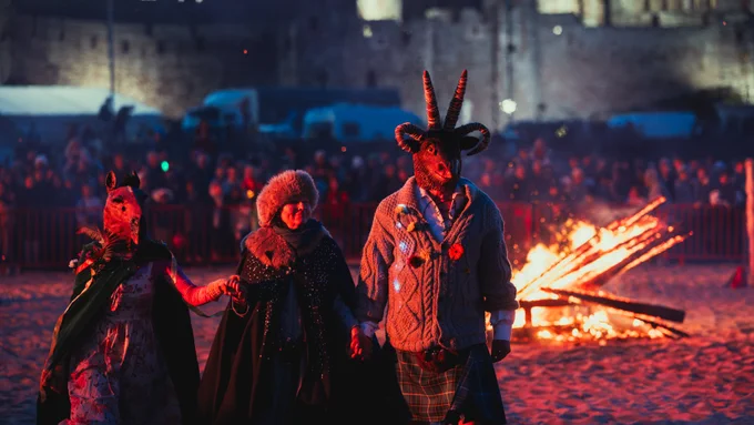 Costumed characters stand together on a beach with a small bonfire and crowd of people in the background