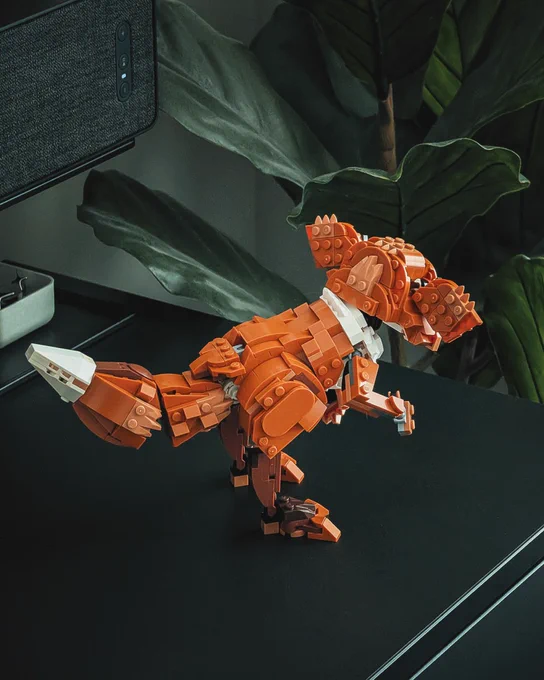An indoor setting with a black shelf, speaker, and artificial plant displays a LEGO brick-built Bogling, a short, furry creature with a long, fluffy tail, two-toed,bird-like feet, and two large, fluffy ears that are lowered in fear. The model balances on two legs.