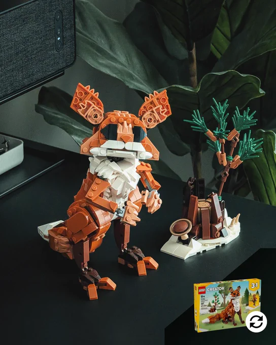 An indoor setting with a black shelf, speaker, and artificial plant displays a LEGO brick-built Bogling, a short, furry creature with a long, fluffy tail, two-toed,bird-like feet, two small hands collapsed across its chest, a large head with very large ears, and two giant, unsettlingly black almond-shaped eyes. Next to the Bogling is a LEGO brick-built tree stump on a snowy diorama base, with alien mushroom and alien tree growing out of it.
