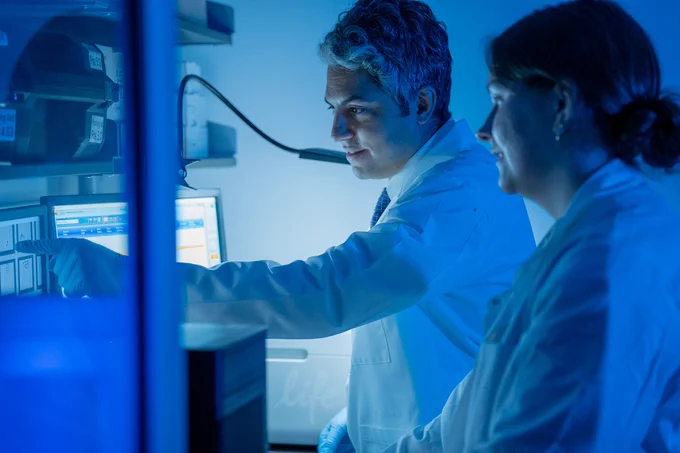Two scientists in lab coats are engaged in a discussion while examining data on a computer screen. The environment is dimly lit with blue tones, suggesting a high-tech laboratory setting.