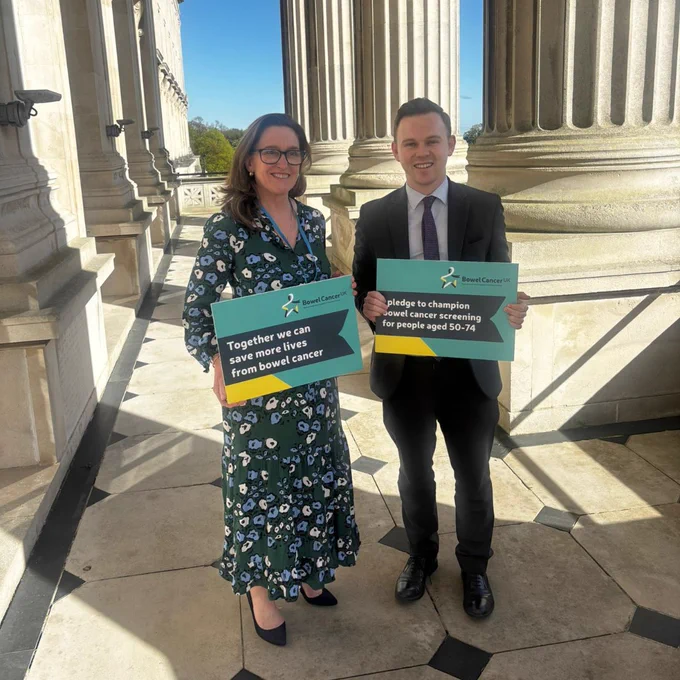 Bowel Cancer UK's Chief Executive Genevieve Edwards stood next to Eóin Tennyson MLA, Deputy Leader of the Alliance Party, outside of Stormont. They're both holding pledge cards calling for Northern Ireland to lower the screening age to 50. 
