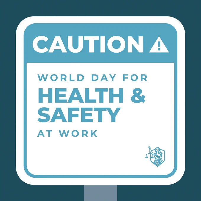 Sign with bold text reading "Caution: World Day for Health & Safety at Work" with an exclamation mark symbol. The background is blue, and a small MAJ logo is in the bottom right corner.
