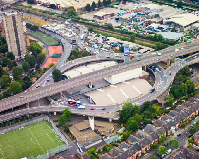 Aerial shot of a highway intersection with cars, buses, and trucks moving on multiple levels. A building and a sports field sit adjacent to the highway, with houses lined with trees on the other side of the road.