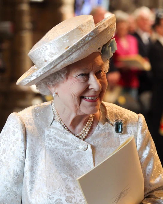 Queen Elizabeth II smiling as she stands in Westminster Abbey wearing a cream hat and coat with an aqua-coloured brooch.