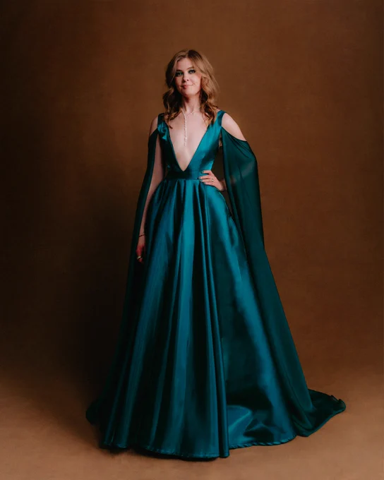 Jennifer English poses with one hand on her hip. She is wearing a long turqouise dress that drapes over her arms and a beaded necklace.