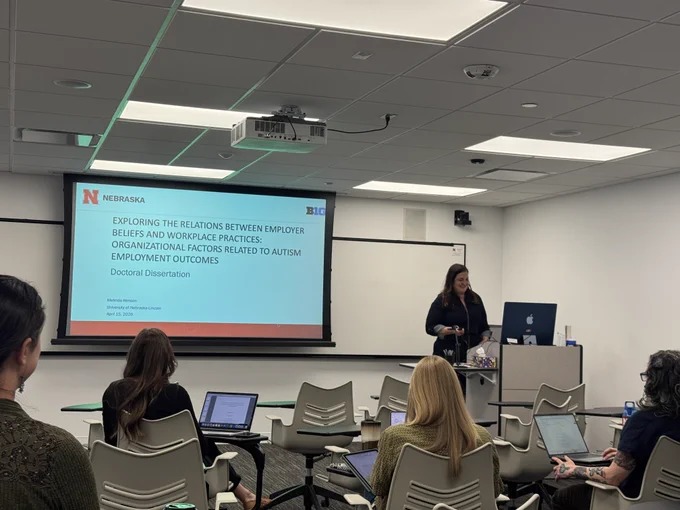 Dr. Melinda Henson defending her dissertation in front of a classroom full of faculty. In the front is a projector with her dissertation title displayed. The room is surrounded by white walls, ceiling tiles, and bright colored LED lights.