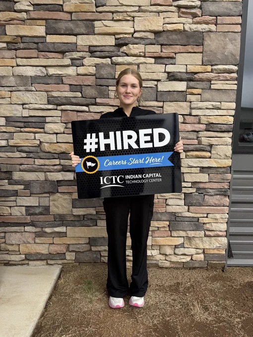 Person in black clothing holding a sign that says "#HIRED" in front of a brick wall.