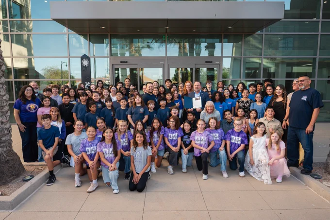 A large group of children, teens, and adults gather outside a building as a male official holds a proclamation document at the center.