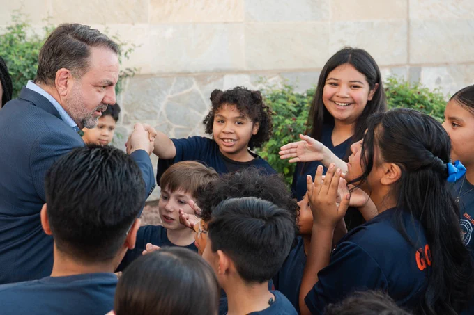A male adult interacts with a group of children outdoors, leaning in as the children smile, gesture, and engage with him.