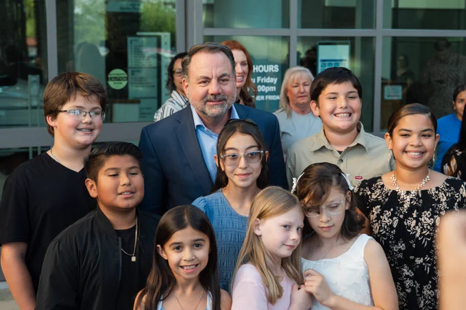 A male adult stands with a group of elementary-aged children, smiling together for a photo outside a building entrance.
