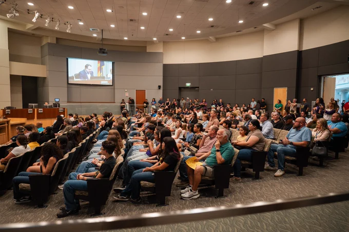 A large audience of men, women, and children sit in rows inside a council chamber, watching a meeting in progress.