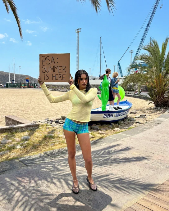 An image of a person wearing a pair of black JUJU JELLIES Maddi Ballerina shoes with a summery outfit whilst standing on a beach front holding a sign that says "PSA SUMMER IS HERE".