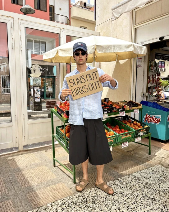 An image of a person wearing a pair of taupe BIRKENSTOCK Arizona sandals with a summery outfit whilst standing in a green grocer holding a sign that says "SUN'S OUT BIRKS OUT".