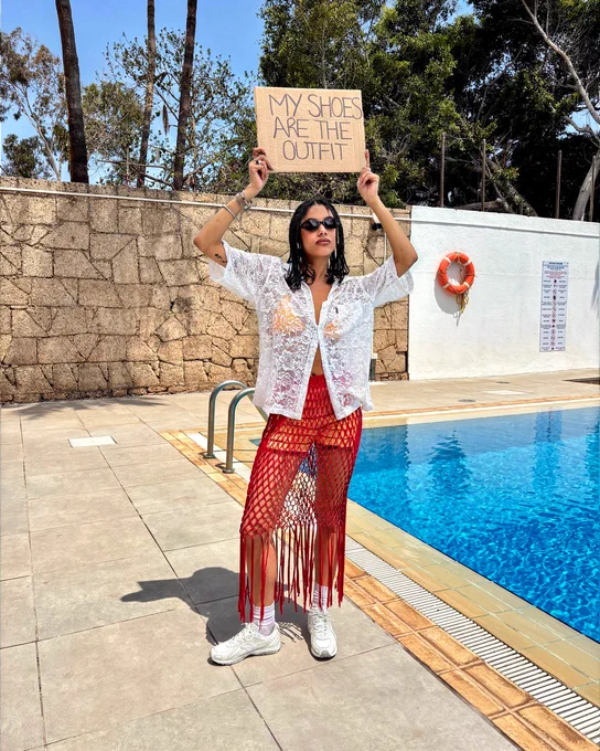An image of a person wearing a pair of cream and pink New Balance 204L trainers with a summery outfit whilst standing next to a pool holding a sign that says "MY SHOES ARE THE OUTFIT".