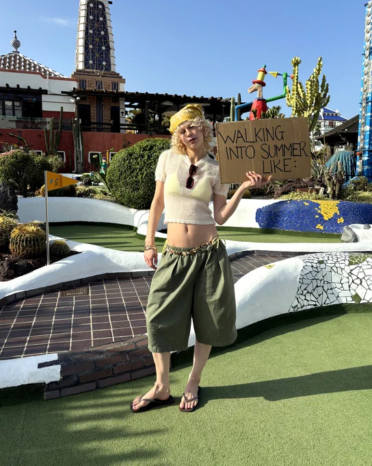 An image of a person wearing a pair of brown Havaianas Slim Square Flip sandals with a summery outfit whilst standing on a mini golf course holding a sign thay says "WALKING INTO SUMMER LIKE".