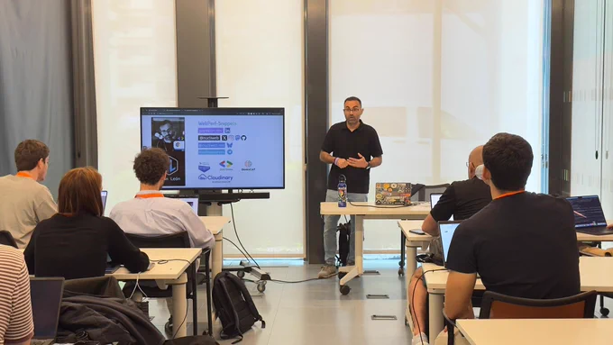 Joan León stands at the front of a classroom giving a presentation to a small group of seated attendees. He gestures while speaking beside a large screen displaying slides about web performance. The audience, some using laptops, face him attentively in a modern, well-lit room with large windows.