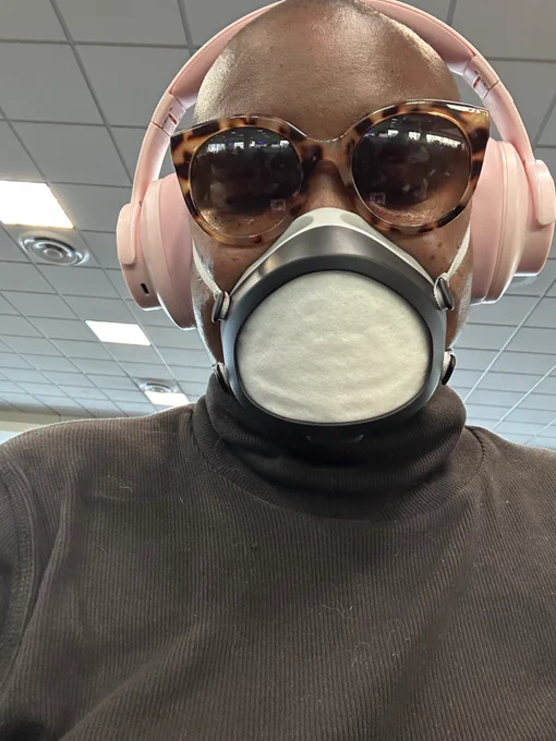 A Black person with a shaved head faces the camera. They are wearing leopard print shades and pink over the ear headphones along with a black Flo mask.