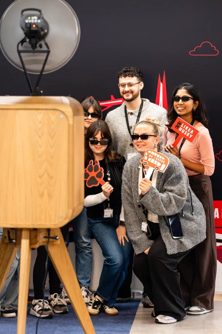 Five people pose at a photo booth with detective-themed props. Two women hold \"NU LONDON\" bus and \"#LIKEAHUSKY\" signs. All are smiling and posing.