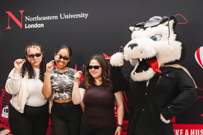Three students wearing sunglasses and holding magnifying glasses pose next to the Northeastern University London husky mascot, Paws, against a black background with the university logo. Paws the mascot is dressed as Sherlock Holmes, with a deer stalker and black jacket.