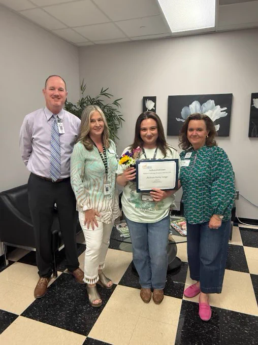 Four people stand in an office-like setting with a checkered floor, smiling for a group photo. In the center, a young woman holds a bouquet of flowers and a certificate that identifies her as the "Midlands Technology Champion of the Year." She is flanked by a man in a lavender dress shirt and tie on the left, and two women on either side. Flower artwork hangs on the wall in the background.