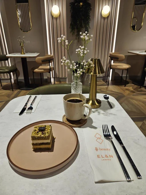 Up market cafe with a square of cake on a white plate on white table. Cutlery and serviettes with a cup of black coffee. Other tables, flowers, lighting and mirrors