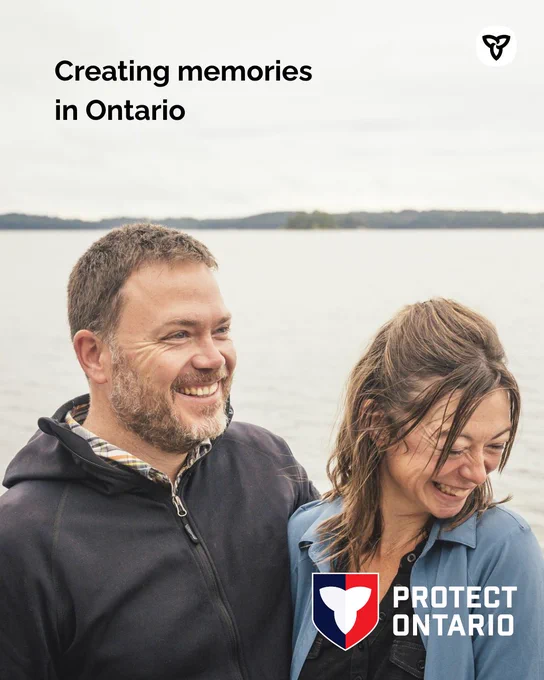 Two people stand side by side near the edge of a lake, with calm water and a distant shoreline visible behind them under an overcast sky. One person wears a dark jacket and the other wears a light blue shirt. Text on the image reads “Creating memories in Ontario,” with the Protect Ontario shield logo displayed in the bottom corner.