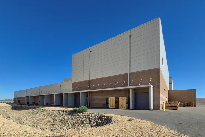 Large industrial warehouse with multiple loading docks under a clear blue sky and gravel foreground.
