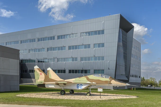 Camouflaged military jet displayed on a circular gravel patch outside a modern gray office building under a partly cloudy sky.